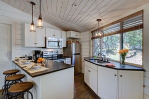 Una cocina con armarios blancos, una isla central con taburetes, un refrigerador de acero inoxidable y una ventana con vista a zonas verdes.