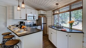 A kitchen with white cabinets, a central island with stools, stainless steel appliances, and a window with a view of trees.