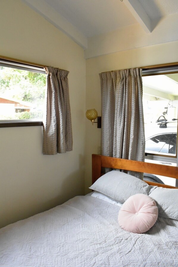A single bed with a wooden headboard, two windows with curtains, and a wall-mounted lamp.