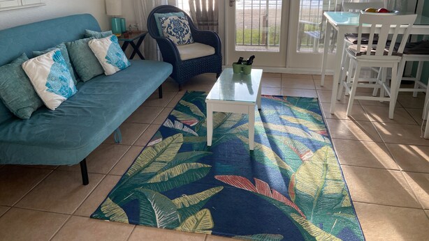 A living room with a blue sofa, a patterned rug, a glass table, and a view of the ocean. - One Bedroom Oceanfront Condo With Room for Six (South Padre Island)