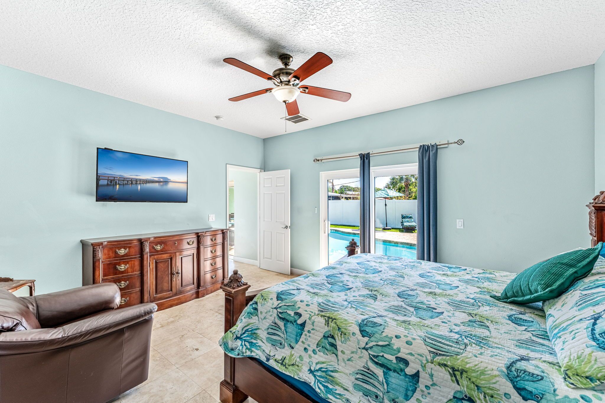 A bedroom with a bed, a ceiling fan, a dresser, a TV, and a view of a pool.