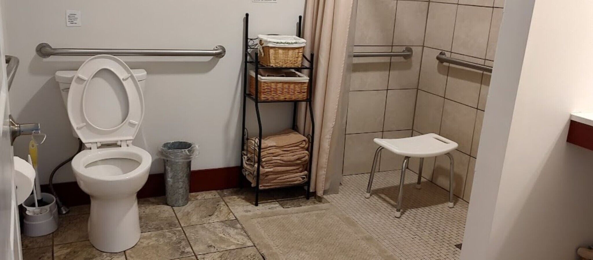 A bathroom with a toilet, a trash can, a shelf with baskets, and a shower area.