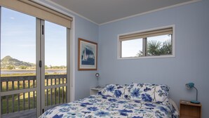 A bedroom with a bed, a nightstand, a framed picture, and a window with a view of a landscape. - Perfect for the golfer or the family beach holiday (Tairua)