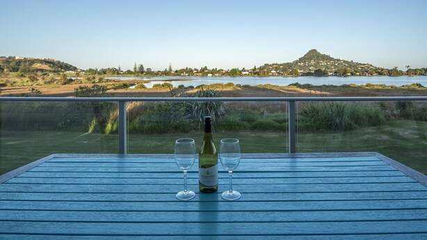 A table with a bottle of wine and two glasses, overlooking a scenic view of a lake and hills. - Perfect for the golfer or the family beach holiday (Tairua)