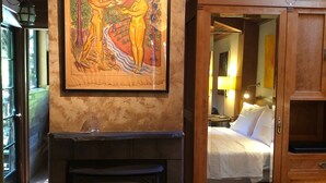 A fireplace with a framed painting above it, a wooden cabinet with a mirror, and a bed with white linens in the background.