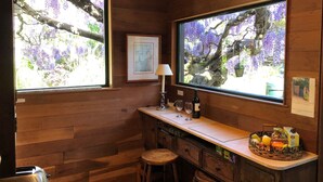 A wooden cabin with two large windows overlooking purple flowers. A small bar area with a lamp, wine bottle, and glasses is visible.