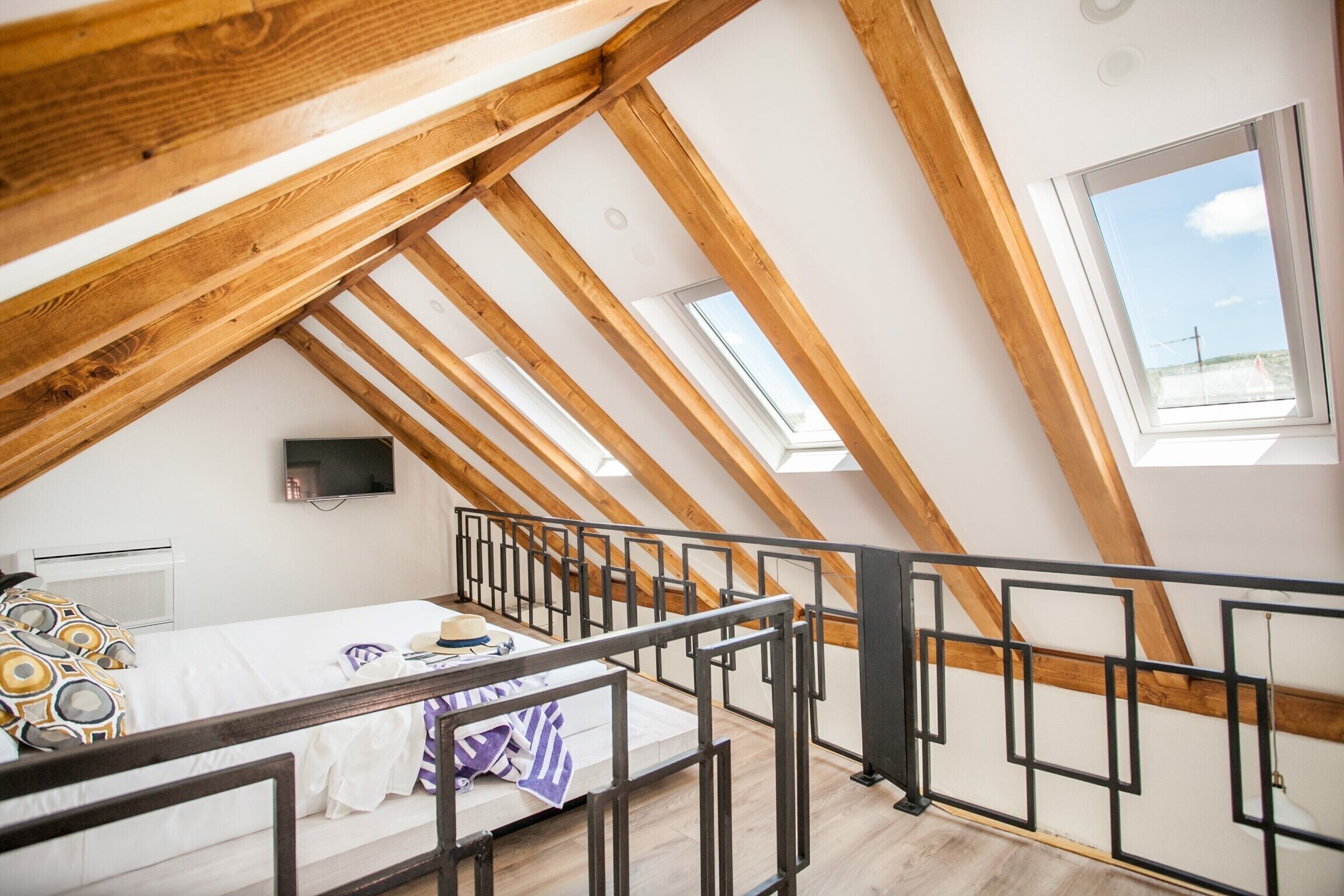 A bedroom with a sloped ceiling, wooden beams, a bed with a white comforter, a TV on the wall, and a window with a view of the sky.