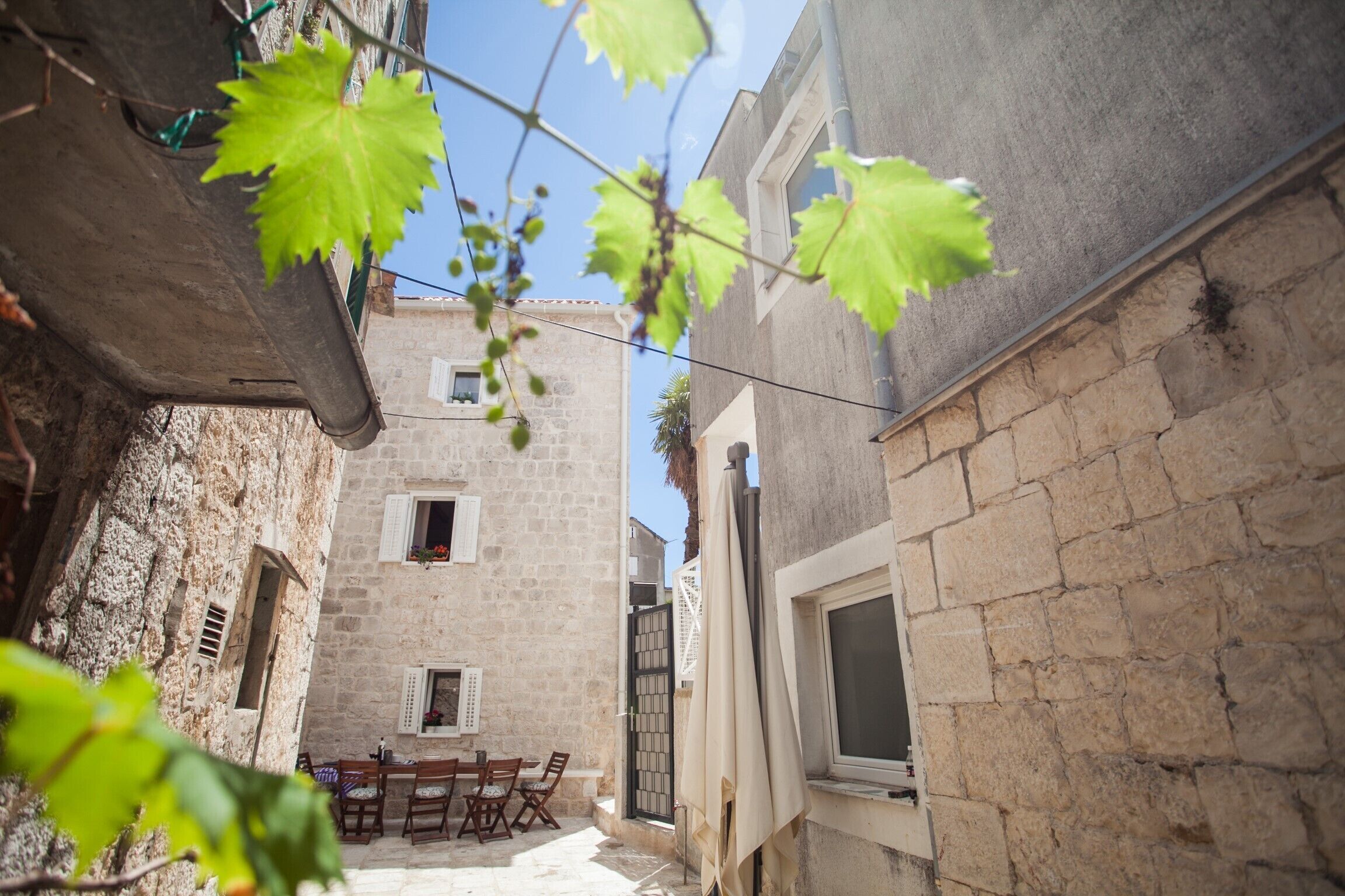 A narrow courtyard with stone buildings, a table with chairs, and a window overlooking the area.