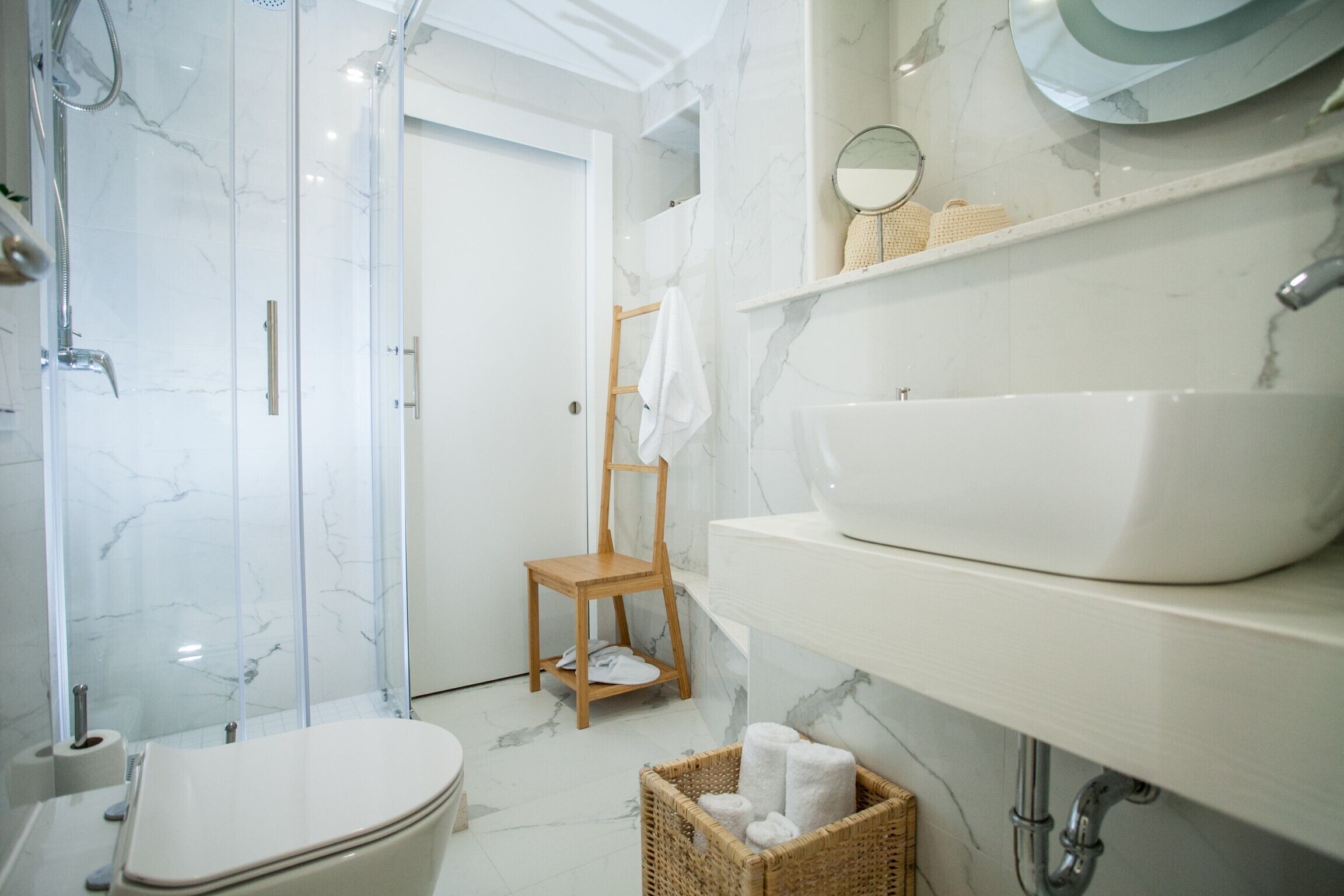Une salle de bain moderne avec une douche à parois de verre, une chaise en bois, un lavabo blanc et un sol carrelé en marbre.