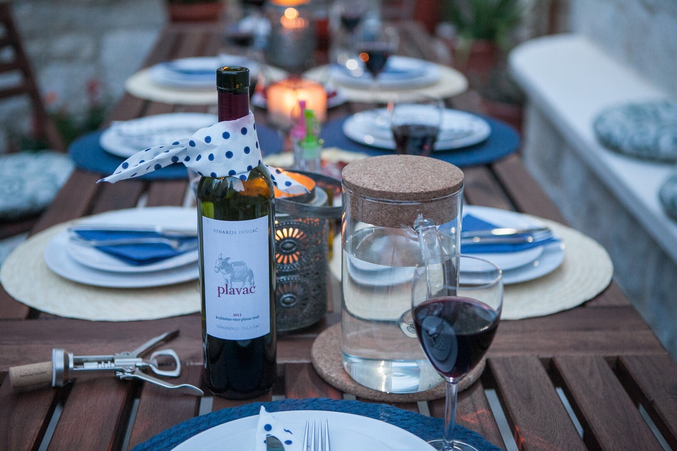 A dining table set with plates, cutlery, a wine bottle, a corkscrew, a glass of red wine, and a candle.