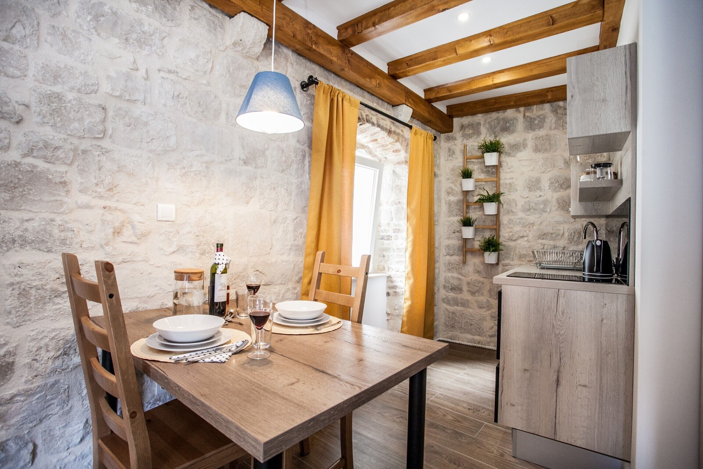 A rustic dining area with a wooden table set for two, a stone wall, and a kitchen with wooden cabinets.