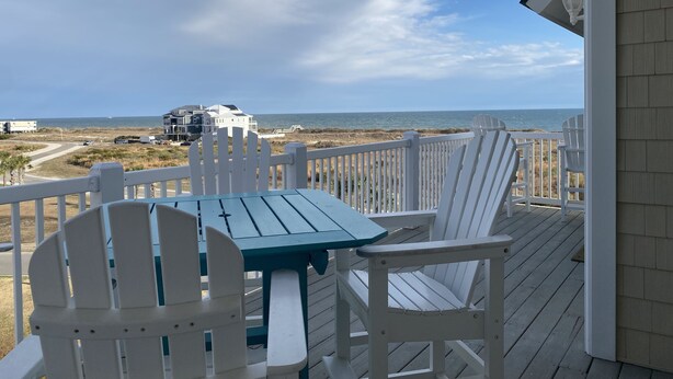 A wooden deck with a table and chairs, overlooking a beach with houses in the distance. - Condo with AMAZING Ocean View! Labor Day Weekend is available and discounted ! (Ocean Isle Beach)