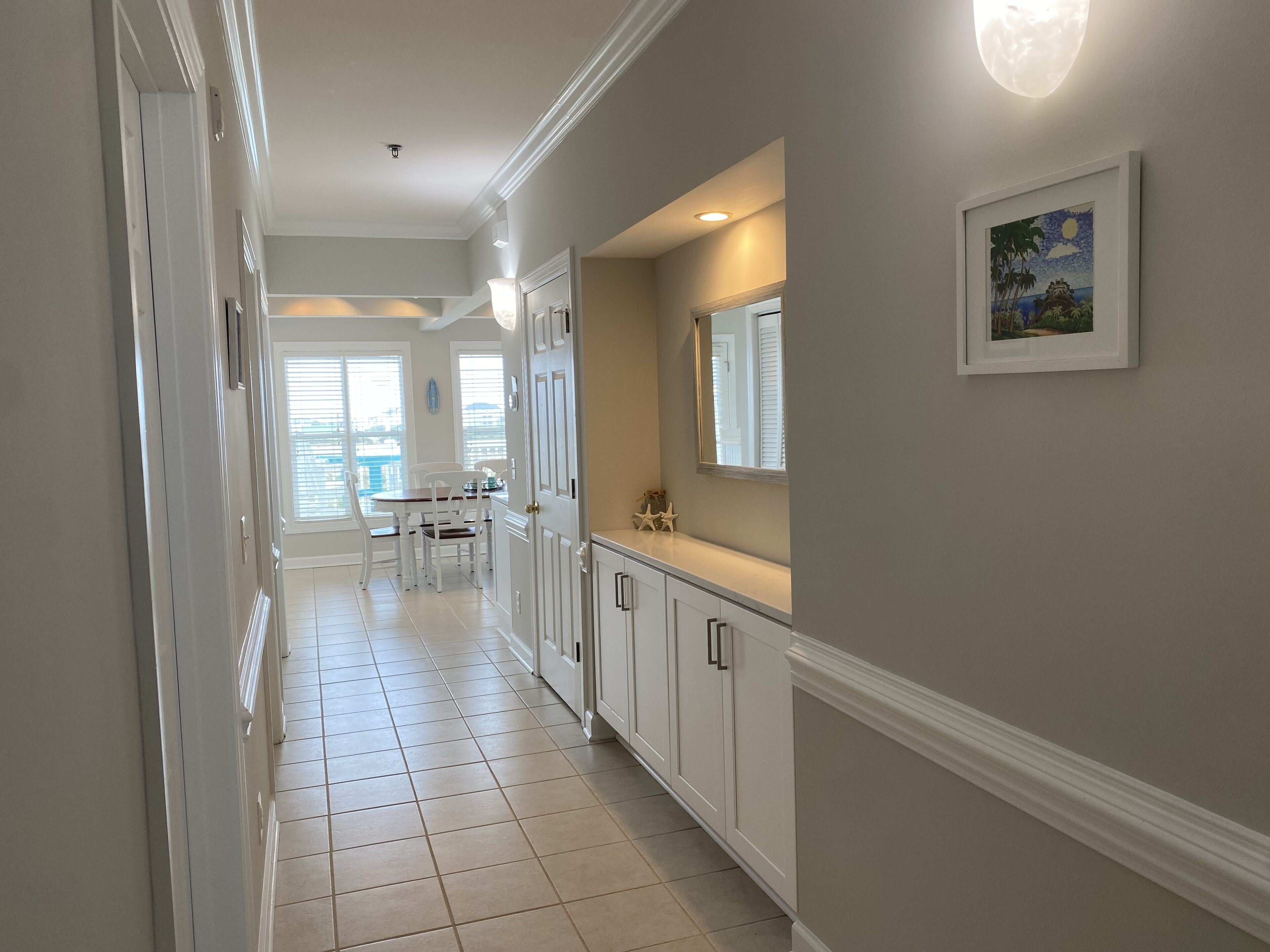 A hallway with white cabinets, a dining area with a table and chairs, and a large mirror on the wall.