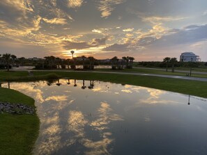 A serene sunset over a pond with reflections of clouds, surrounded by greenery and a distant building. - Condo with AMAZING Ocean View! Labor Day Weekend is available and discounted ! (Ocean Isle Beach)
