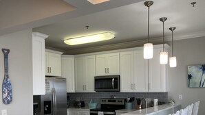 A modern kitchen with white cabinetry, stainless steel appliances, a central island with bar stools, pendant lighting, and a painting on the wall. - Condo with AMAZING Ocean View! Labor Day Weekend is available and discounted ! (Ocean Isle Beach)