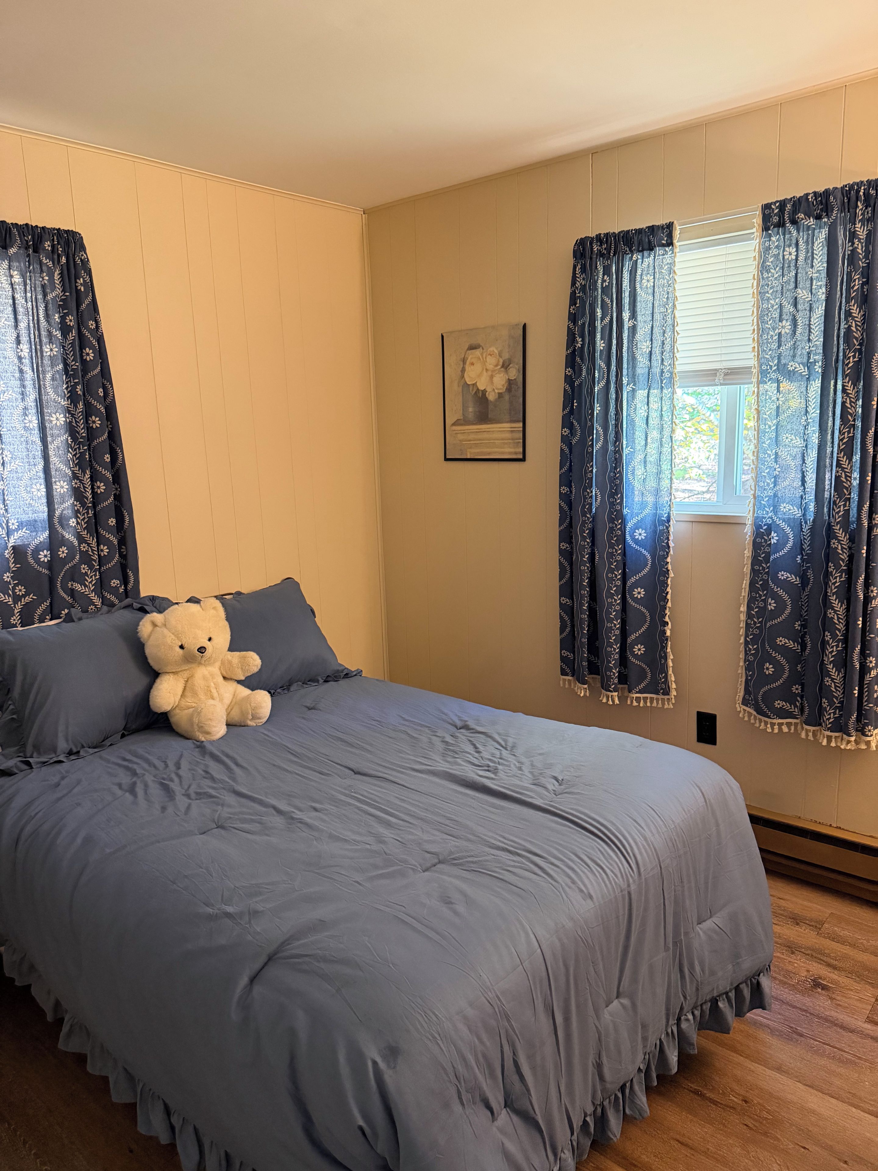 Une chambre avec un grand lit, un ours en peluche posé sur le lit et une fenêtre avec des rideaux.