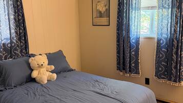 Une chambre avec un grand lit, un ours en peluche posé sur le lit et une fenêtre avec des rideaux.