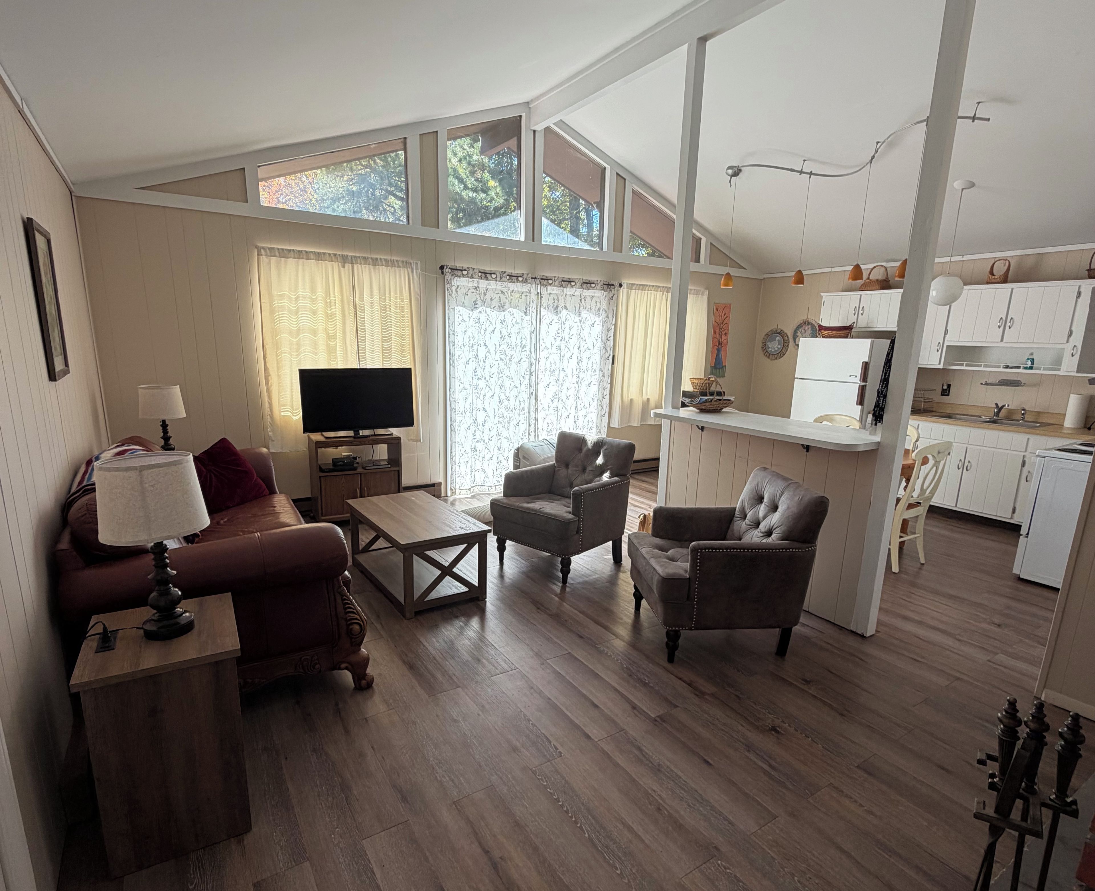 Amplia sala de estar con sofá, sillones, una mesa de centro y televisor. Se observa una zona de cocina con armarios blancos y un refrigerador.