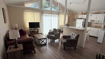 Amplia sala de estar con sofá, sillones, una mesa de centro y televisor. Se observa una zona de cocina con armarios blancos y un refrigerador.