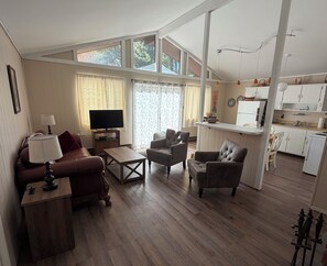 A spacious living area with a sofa, armchairs, a coffee table, and a television. There is a kitchen area with white cabinets and a refrigerator.
