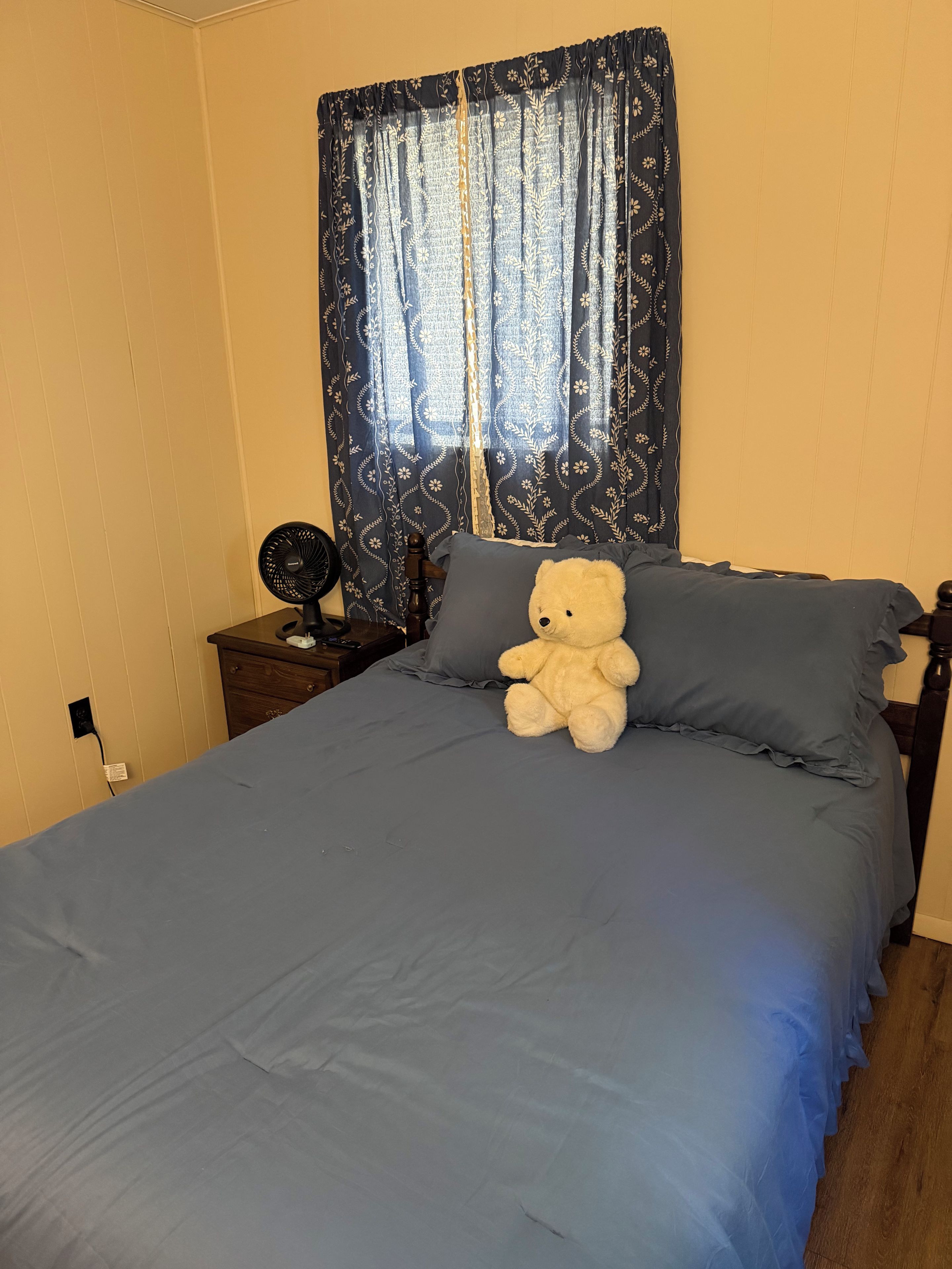 A bed with a blue bedspread, a teddy bear on the pillows, a nightstand with a fan, and a window with patterned curtains.