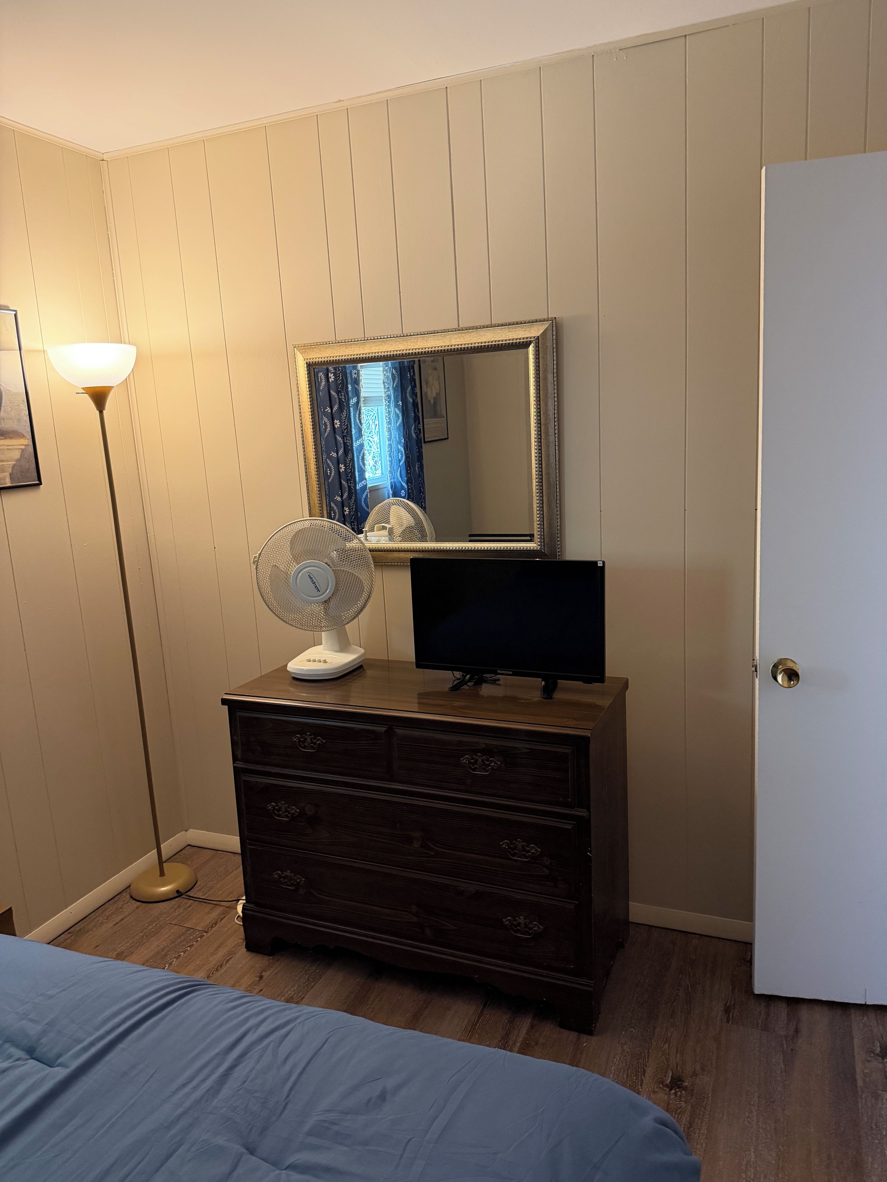 A bedroom with a dresser, a television, a standing fan, and a bed.