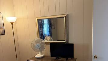 A bedroom with a dresser, a television, a standing fan, and a bed.
