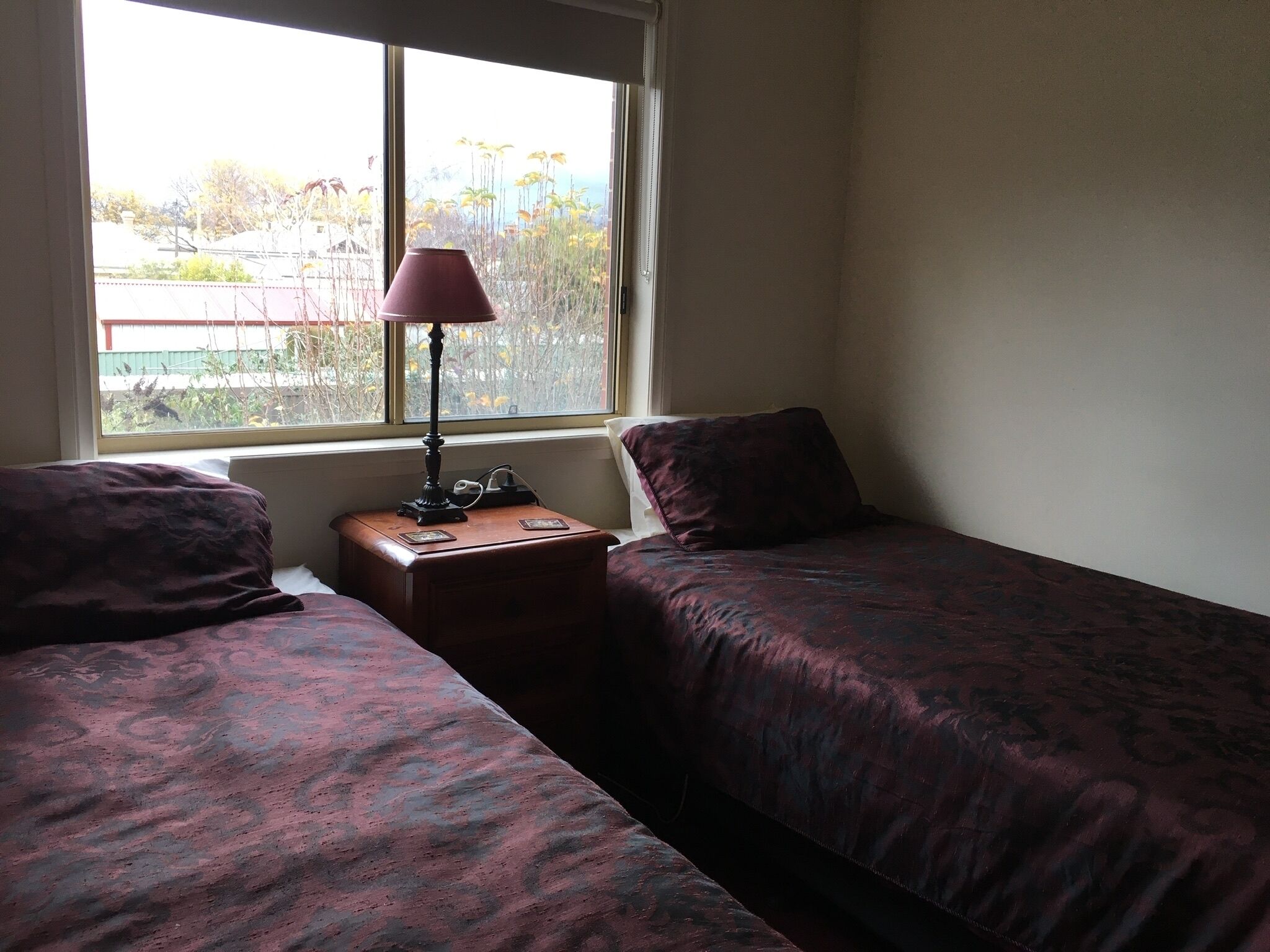 A room with two beds, a nightstand with a lamp, and a window with a view of trees and buildings.