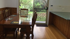 A dining area with a wooden table and chairs, a view through a glass door to a garden, and a clock on the wall.