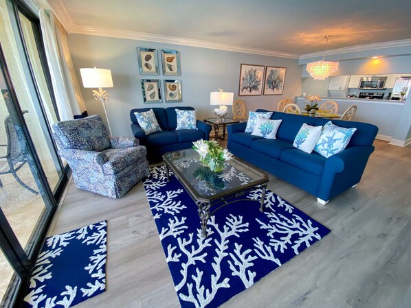 Una sala de estar con un sofá azul, un sillón estampado y una mesa de centro de vidrio con un centro de mesa floral.
