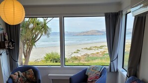 Une pièce avec deux fauteuils bleus, un canapé bordeaux et une vue sur la plage et l’océan, grâce à de grandes fenêtres.