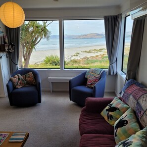 Une pièce avec deux fauteuils bleus, un canapé bordeaux et une vue sur la plage et l’océan, grâce à de grandes fenêtres.