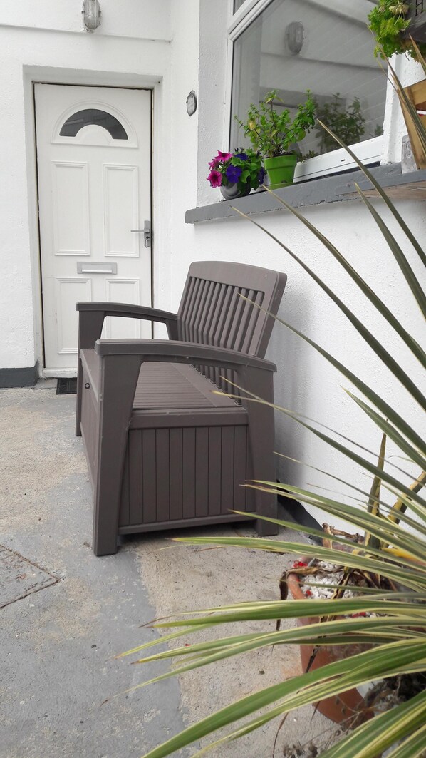 Une terrasse avec une chaise marron et une petite table, une porte blanche et une fenêtre ornée d’un pot de fleurs.