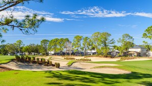 Een golfbaan met zandbunkers, bomen en moderne gebouwen op de achtergrond.