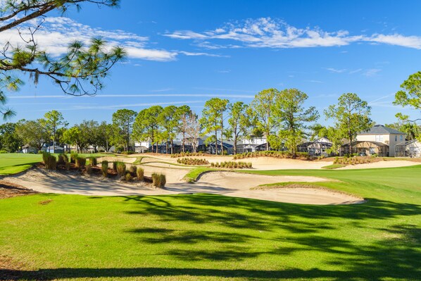 A golf course with sand bunkers, trees, and modern buildings in the background.