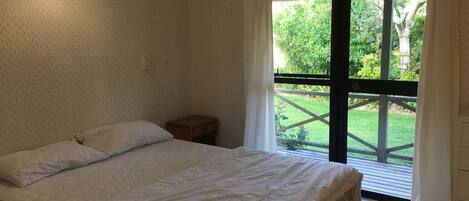 Un dormitorio con cama, mesita de noche, ventana con vistas a zonas verdes y una lámpara en el techo.