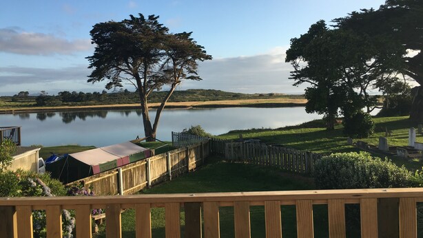 A lakeside view with a wooden deck, a hammock, and a fenced area with a vehicle. - Riverfront Cottage Waipu (Waipu)