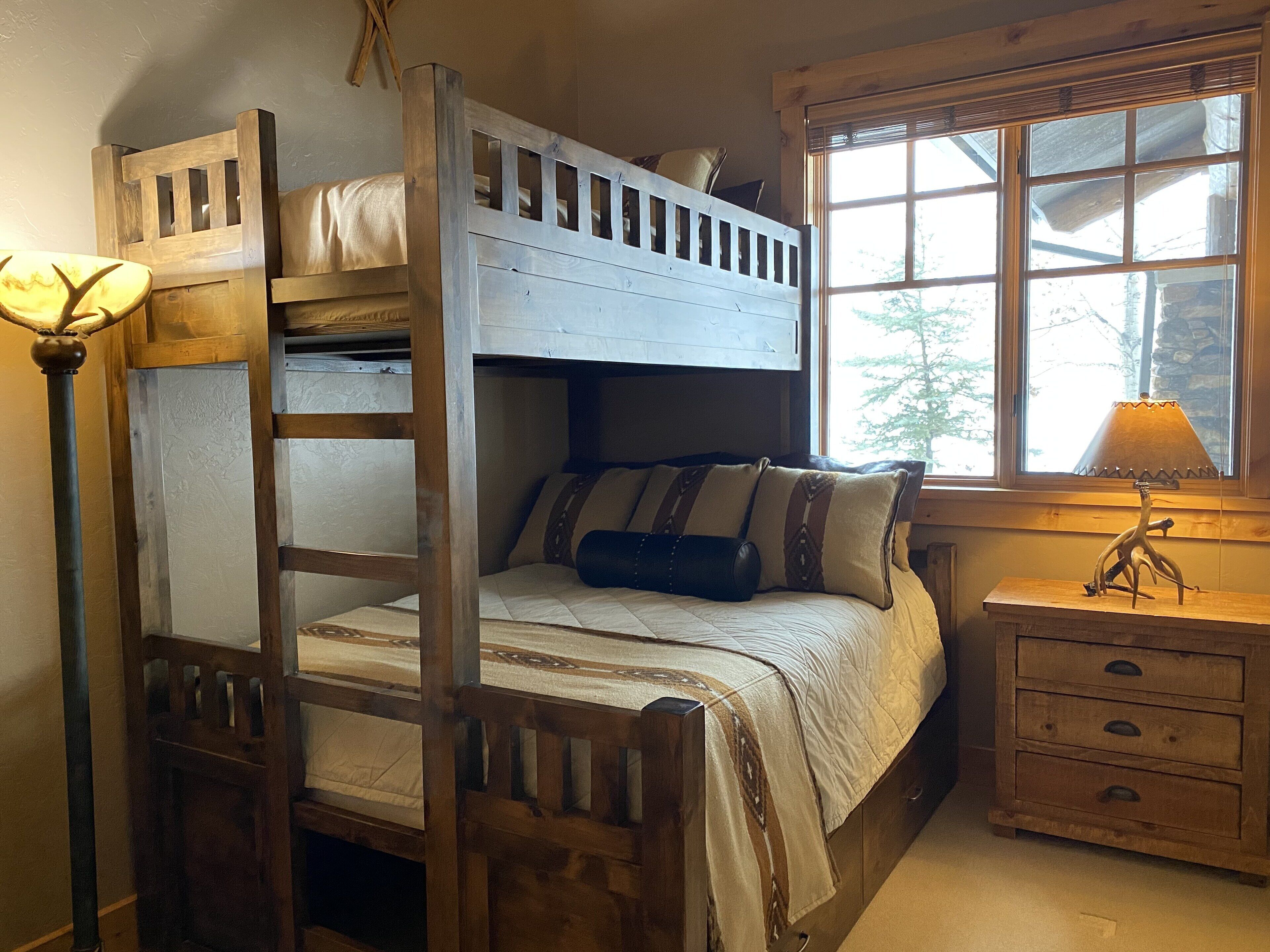 A bunk bed with a ladder, a bedside table with a lamp, and a window with a view of trees.