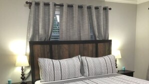 A neatly made bed with a white comforter and pillows, flanked by two bedside tables with lamps, in a room with a dark wooden headboard and a view of a staircase through a window with grey curtains. - Private 4BR Heated Pool & Spa Home • Pet Friendly • Near Beaches (Margate)