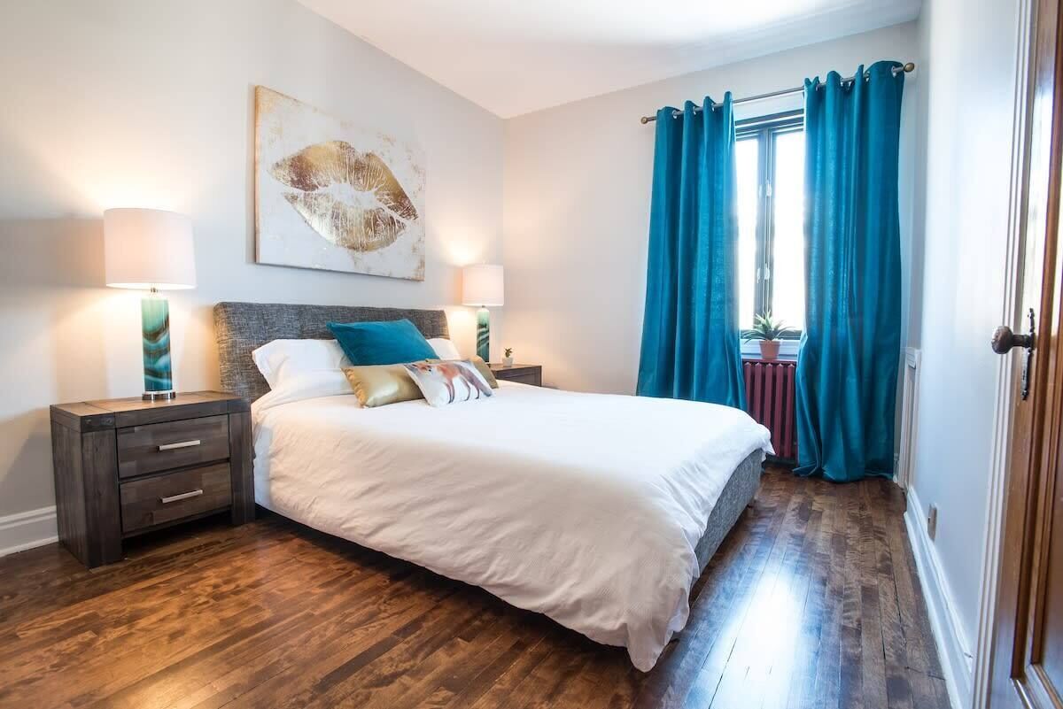 Un dormitorio con una cama grande, dos mesitas de noche, una mesita auxiliar, una ventana con cortinas y un radiador.