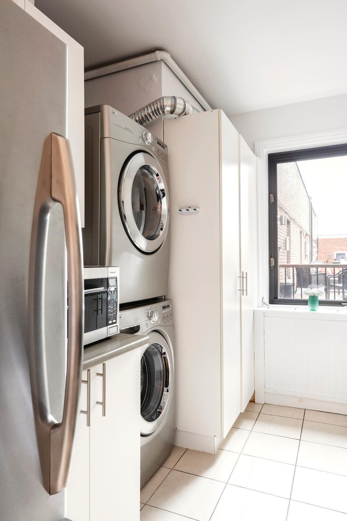 A compact kitchen with a built-in washer and dryer, a stainless steel refrigerator, and a window with a view of the street.