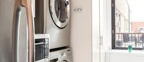 A compact kitchen with a built-in washer and dryer, a stainless steel refrigerator, and a window with a view of the street.