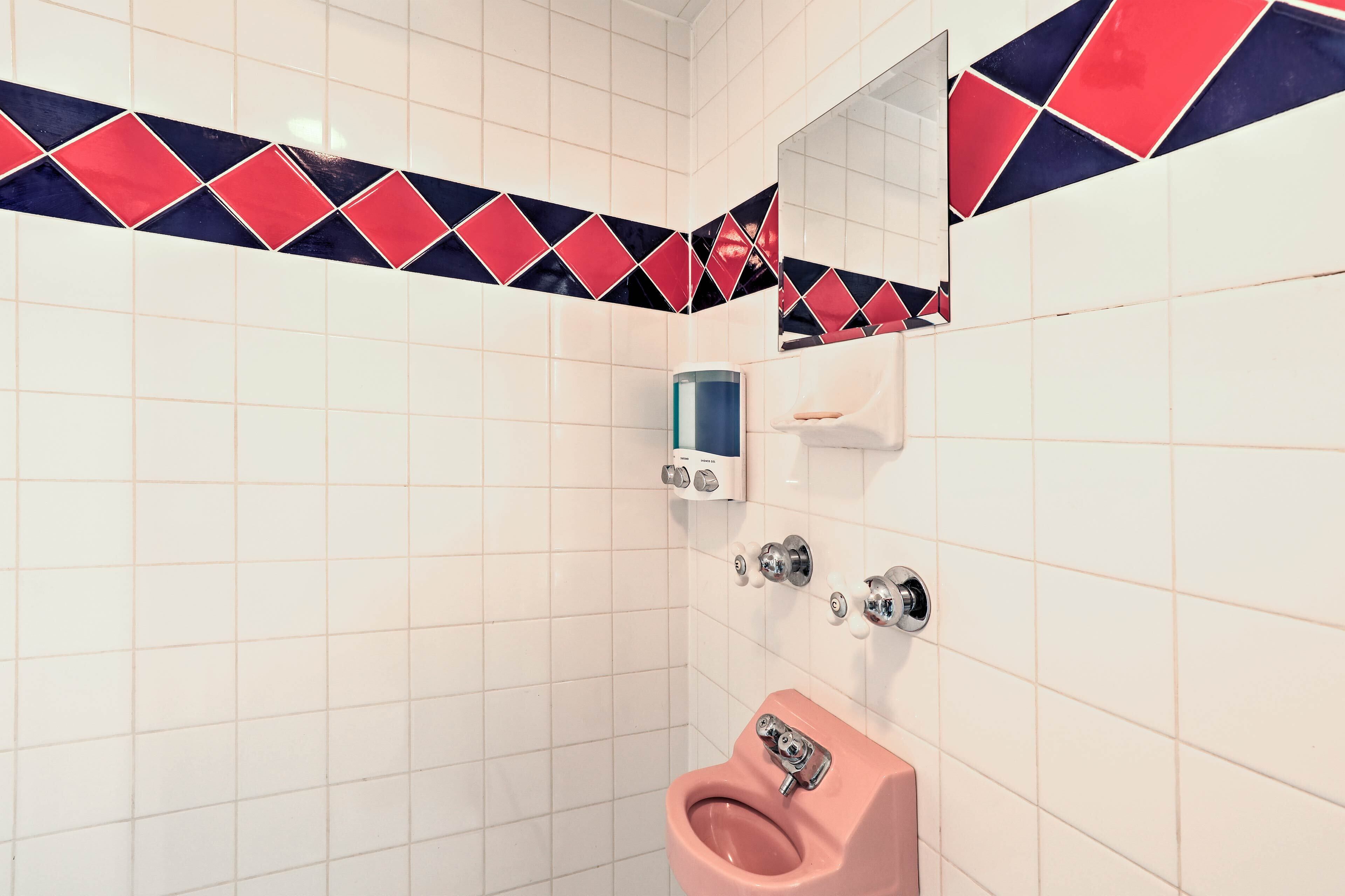 Ein Badezimmer mit weißen Fliesen, einem rosa Urinal, einem Handtrockner und einem Spiegel mit einem roten und blauen Diamantmuster als Rückwand.