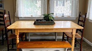 Een eetgedeelte met een houten tafel en bank, twee ramen met witte gordijnen, en een plant op tafel.