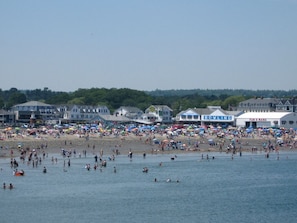 Ein Strand mit badenden und sonnenden Menschen, im Hintergrund sind Gebäude zu sehen.