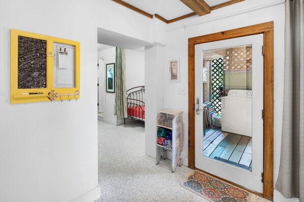 A hallway with a yellow-framed chalkboard, a metal bed frame, a small wooden shelf with folded clothes, and a patterned rug leading to a door with a wooden frame.