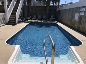 Piscina retangular com uma escada de metal em primeiro plano, cercada por um muro de madeira e palmeiras.