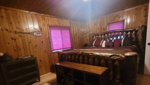 Une pièce chaleureuse aux murs en bois, avec un lit doté d’une tête de lit, un banc, une commode et une fenêtre avec des stores.