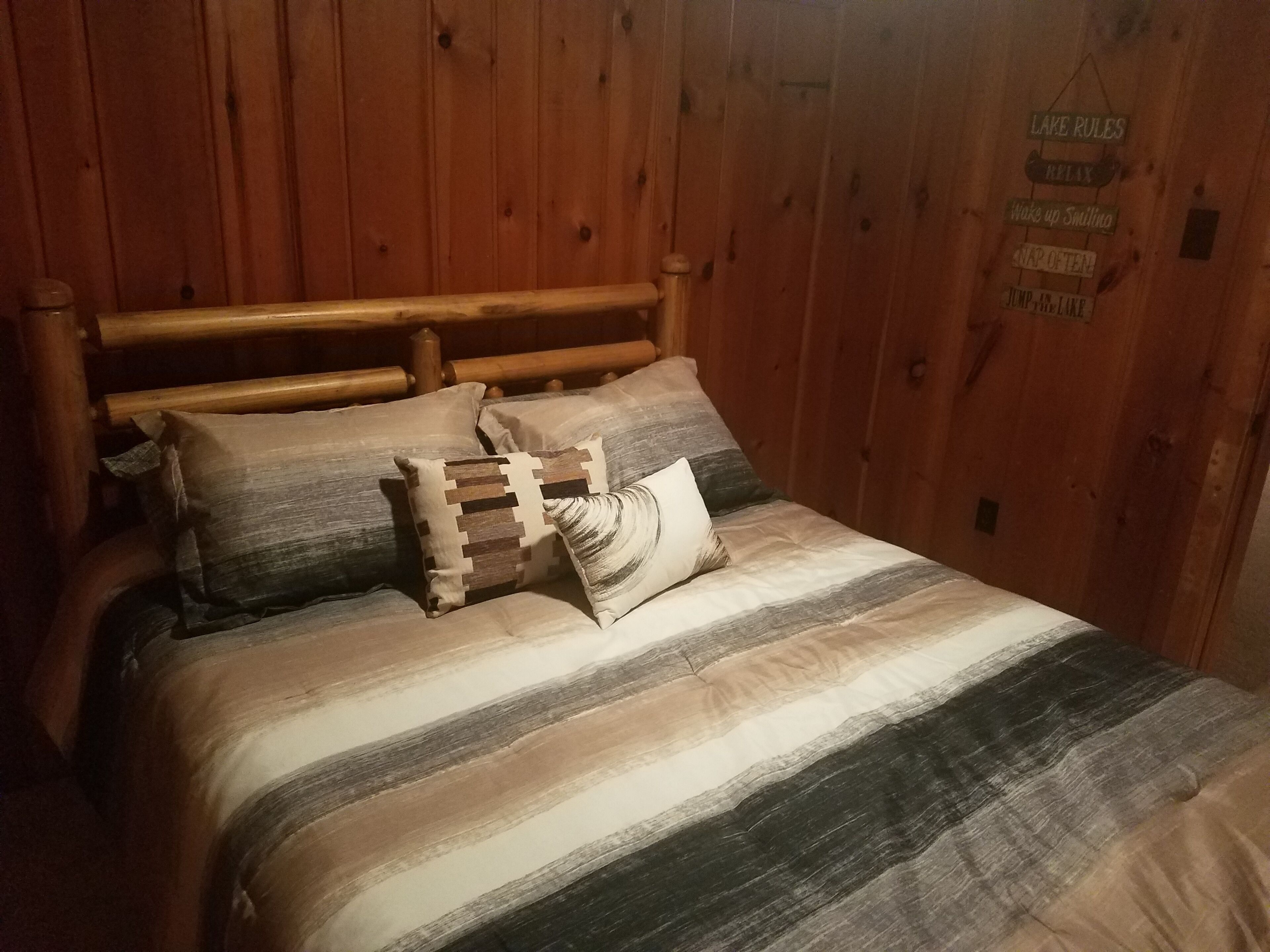 A wooden bed with a striped blanket and pillows, a wooden headboard, and a sign on the wall reading "LAKE RULES".
