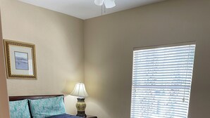 Ein Schlafzimmer mit Bett, Nachttisch, Lampe, einem Fenster mit Jalousien und einem gerahmten Bild an der Wand.